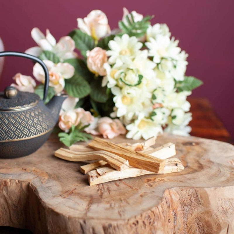 Palo Santo wood on a wooden surface with a teapot and flowers in the background, symbolizing its ritual use.